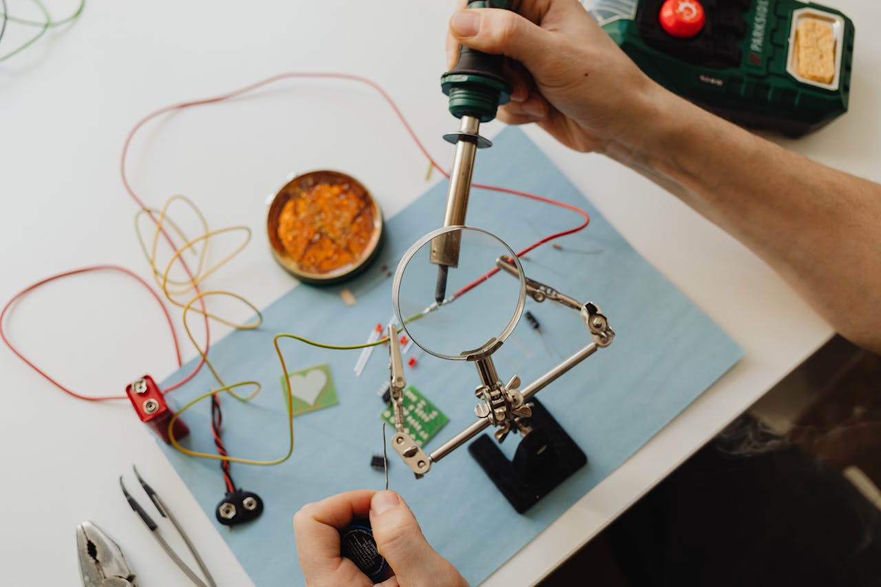 Hands soldering a circuit board using a magnifying glass and tools, from a high angle.